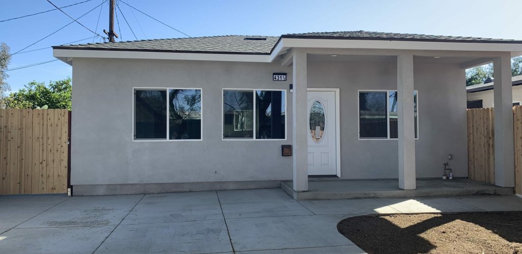 A completed ADU with concrete laid out nicely. The ADU exterior has a light gray tone with a white door. There are two windows on the left of the door and a window on the right of the door. The entry way is also elevated. The roofing is a dark gray.